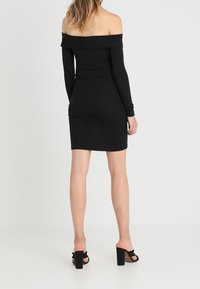 Černé žebrované šaty bez ramínek s dlouhými rukávy, s těsným střihem a délkou do poloviny stehen, v kombinaci s černými sandály na blokovém podpatku.