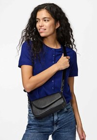 Borsa a tracolla nera con patta curvata, realizzata in materiale testurizzato. Il modello indossa un top blu e jeans di denim mentre tiene la borsa.