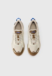 Beigā un brūnā Asics skriešanas apavu pāris ar ziliem papēžu cilpiņām, skatīts no augšas uz vienkārša balta fona.