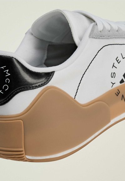 White sneaker with a black heel detail, gray suede accents, and a tan rubber sole. Features a logo on the side and textured materials.
