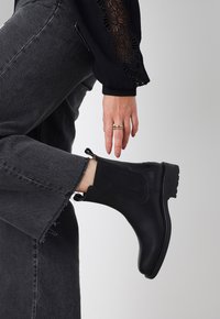 Botín negro hecho de cuero y textil, con paneles laterales elásticos y una suela texturizada. Llevado con jeans gris oscuro.