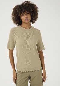 Kurzärmliger Pullover aus Strick in hellem Beige mit strukturiertem, offenem Gewebe, gewelltem Ausschnitt und Saum. Kombiniert mit khakifarbenen Cargo-Hosen.