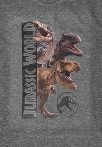Tre dinosauri ruggenti dietro il testo verticale "Jurassic World" e un logo circolare di un dinosauro su uno sfondo di tessuto grigio.