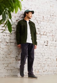 Camicia verde a maniche lunghe, t-shirt bianca, pantaloni grigi in corduroy, scarpe grigie, davanti a un muro di mattoni bianchi testurizzati con foglie verdi nel campo visivo.