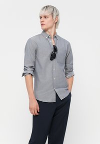 Grauhemd mit Knöpfen und Tasche, hochgekrempelten Ärmeln und leichtem Stoff. Kombiniert mit dunklen Hosen und Sonnenbrille, die auf dem Hemd liegt.