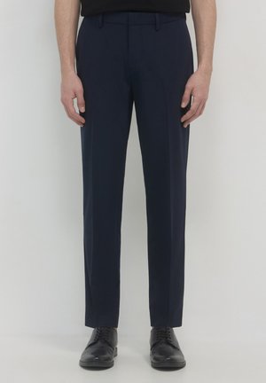 Mann in maßgeschneiderten navyblauen Anzughosen und schwarzen Schnürschuhen, der vor einem schlichten hellen Hintergrund steht.