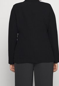 Blazer negro con un diseño estructurado, mangas largas y un acabado de tela suave. La parte posterior presenta costuras ajustadas y un cuello con muesca.