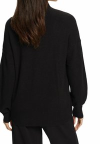 Femme aux longs cheveux bruns portant un pull noir ample et un pantalon noir, vue de dos sur un fond blanc.