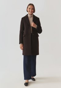 Bruine wollen jas met gekartelde kraag, twee voorzakken en een enkele knoopsluiting. Draagt over een lichte coltrui en donkere wijde jeans.