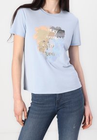 Camiseta de manga corta azul claro con un diseño abstracto colorido y el texto "Sé fiel a ti mismo" en plata. Tejido suave, corte relajado.