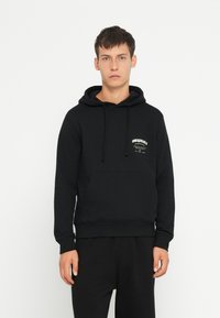 Haut à capuche noir en tissu doux, avec une poche avant, un cordon de serrage à la capuche et un texte graphique blanc sur la poitrine. Design simple, coupe décontractée.
