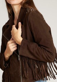 Brązowa zamszowa kurtka z frędzlami na dolnym obwodzie i ramionach. Zawiera srebrne ćwieki oraz zamek błyskawiczny z przodu.