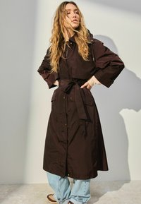 Brun, langærmet trenchcoat med bælte i taljen, trykknapper og påsyede lommer. Kombineret med lyseblå, løstsiddende jeans.