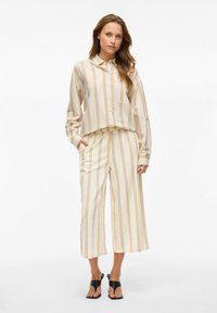 Conjunto ligero de dos piezas a rayas en crema con rayas verticales doradas y marrones. Presenta un cuello, cierre de botones en el frontal y pantalones cortos; se lleva con sandalias negras.