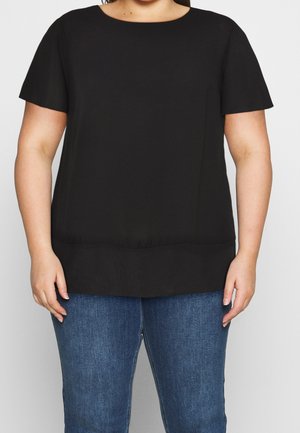 Femme portant un haut noir uni à manches courtes associé à un jean en denim bleu, debout devant un fond clair uni.