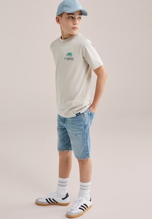 Chlapec oblečený v béžovom tričku s nápisom „Let's Explore“, rifľových šortkách, bielych ponožkách s pásikmi, bielych teniskách a svetlomodrej šiltovke, stojaci s rukami v vreckách.