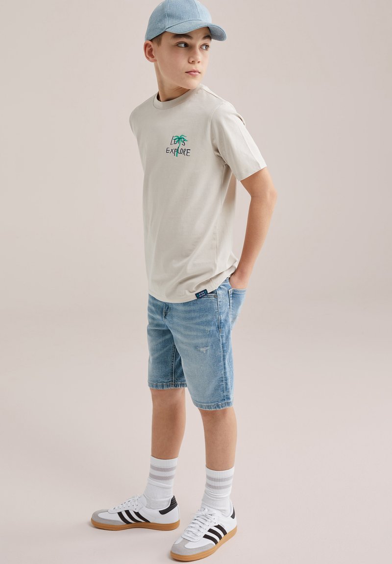 Ragazzo che indossa una maglietta beige con la scritta "Let's Explore", pantaloncini di jeans, calzini bianchi a righe, scarpe da ginnastica bianche e un berretto azzurro, in piedi con le mani nelle tasche.