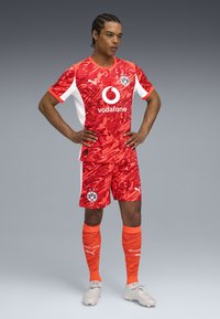 Kit de football à motif camouflage rouge avec des accents blancs, comportant un logo circulaire Vodafone et un écusson BVB. Une paire de chaussettes et de crampons orange vif.