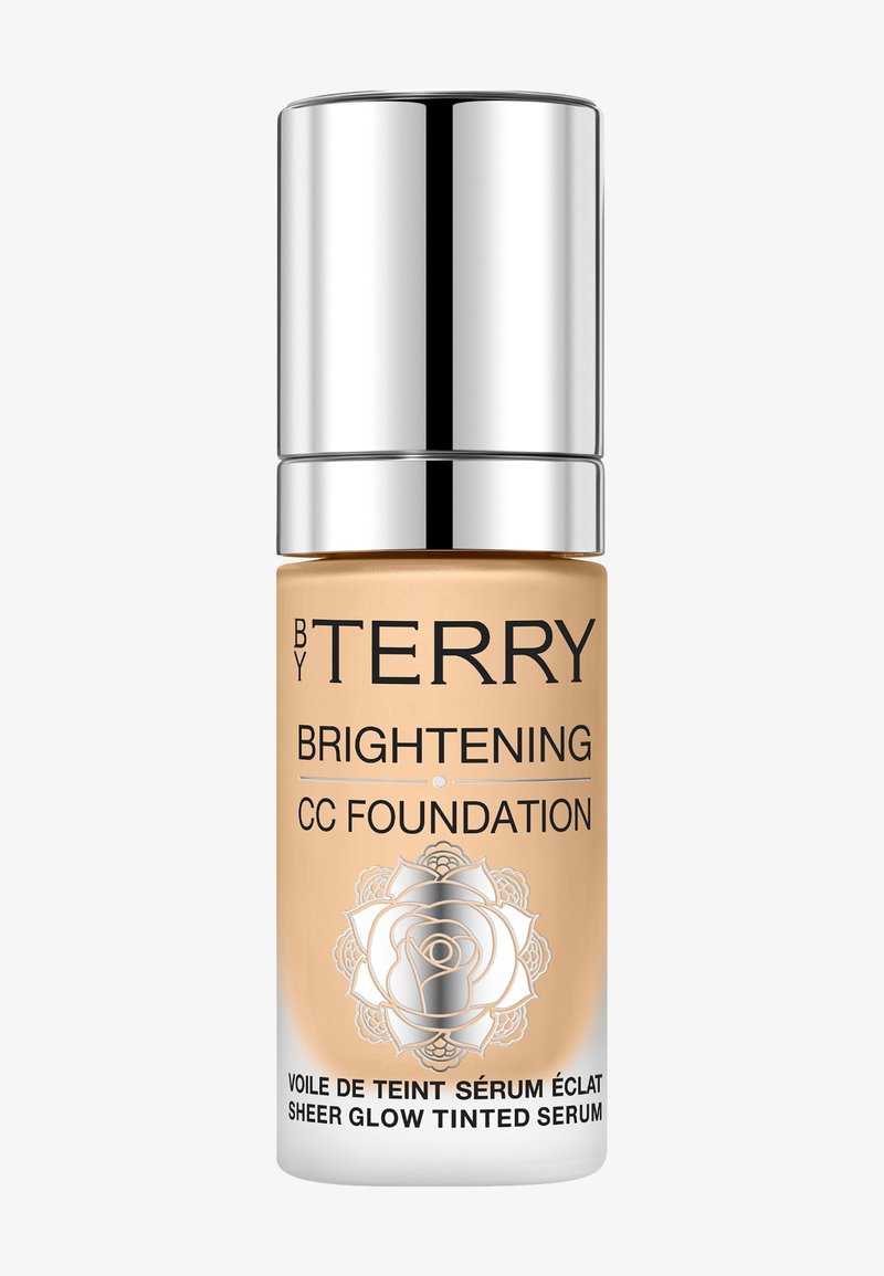 Fond de teint CC dans une bouteille en verre transparente avec un capuchon argenté métallique. Couleur beige clair avec un motif floral sur l'étiquette avant.