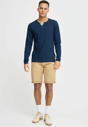 Marineblauwe henley shirt met lange mouwen en een knoopsluiting, gecombineerd met beige shorts, witte crew sokken en lichte sportschoenen met bruine accenten.