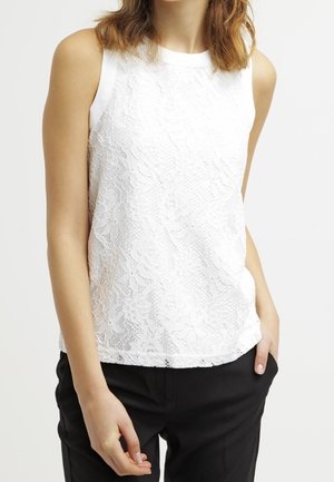 Femme portant un haut en dentelle blanche sans manches et un pantalon noir, une main dans la poche et l'autre détendue le long du corps.