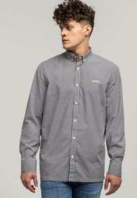 Czarno-biała koszula w kratę z guzikami, z kołnierzykiem, posiadająca długie rękawy i subtelne logo na piersi. Materiał bawełniany, gładka tekstura.