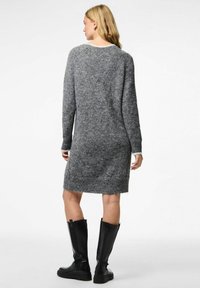 Donna con capelli biondi che indossa un vestito grigio a maglia a maniche lunghe e stivali neri fino al ginocchio, in piedi con la schiena rivolta verso la camera su uno sfondo bianco.