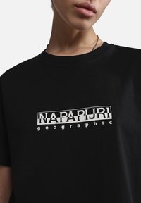 Person iført sort T-shirt med hvid "NAPAPIJRI geographic" logo på brystet, synligt fra nakken til den øverste del af torsoen.