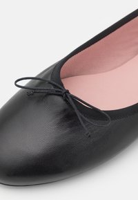 Ballerine en cuir noir avec un nœud sur l’orteil arrondi et une doublure intérieure en tissu rose.