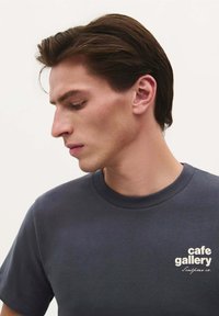 Tricou gri închis din bumbac, cu guler rotund și mâneci scurte. Text imprimat "cafe gallery" în alb pe partea stângă a pieptului.