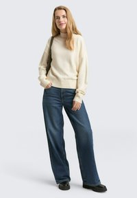 Kremowy sweter z dzianiny z wysokim kołnierzem, zestawiony z szerokimi niebieskimi dżinsami o kroju szerokich nogawek. Modelka ma długie blond włosy i trzyma zieloną torbę.