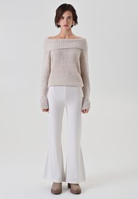 Maglione beige a spalle scoperte in maglia con un ampio scollo a coste, abbinato a pantaloni bianchi aderenti a zampa d'elefante e stivali piattaforma marroni chunky.