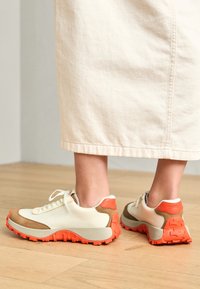 Sneakers con una toma in tela crema, dettagli in suede marrone e suole in gomma arancione brillante. Presentano lacci tondi e un tallone rinforzato.