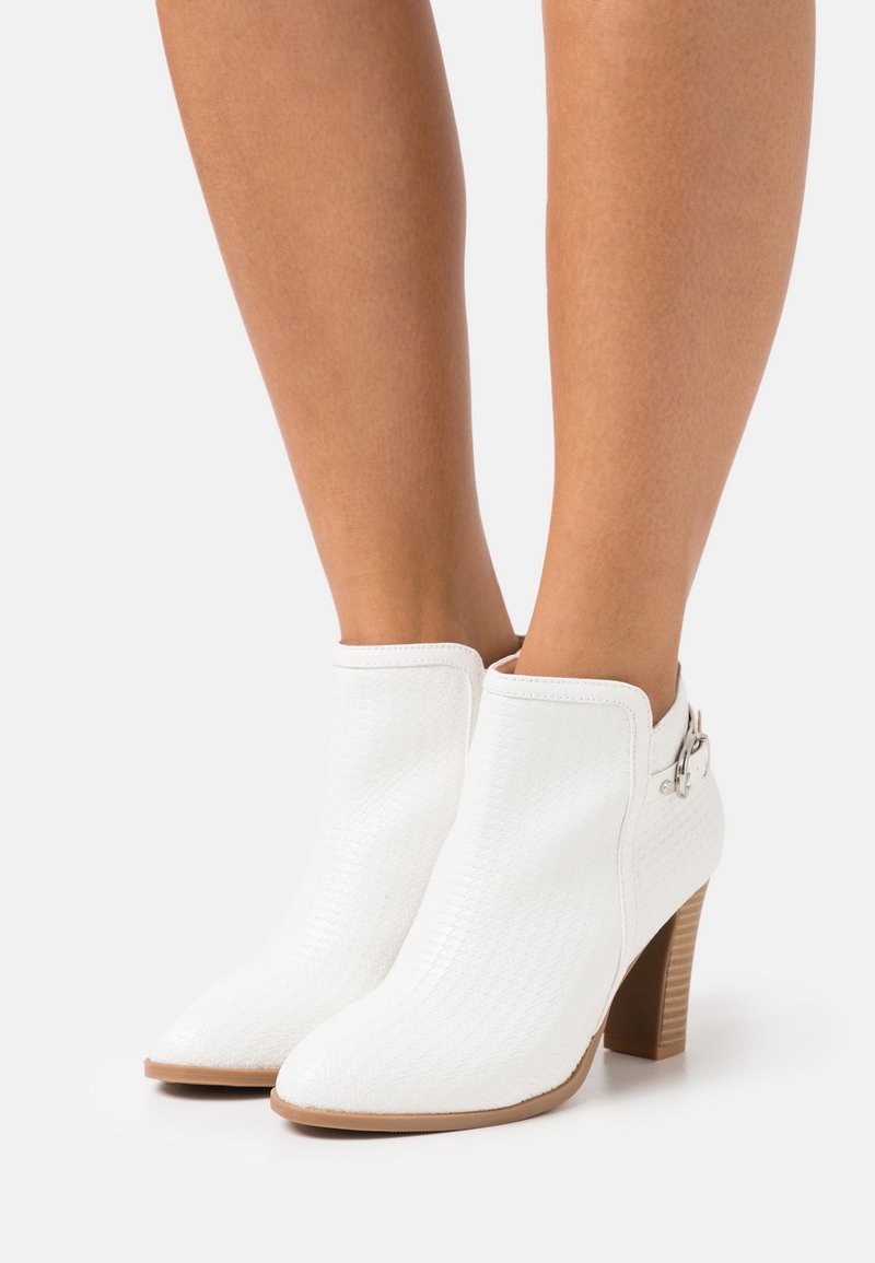 Bottes blanches texturées à cheville avec talons blocs et détail de boucle latérale portées sur des jambes nues sur un fond uni.
