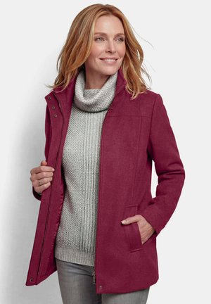 Frau trägt einen kastanienbraunen Mantel über einem grauen Rollkragenpullover, mit einer Hand in der Manteltasche, lächelnd und leicht zur Seite schauend.