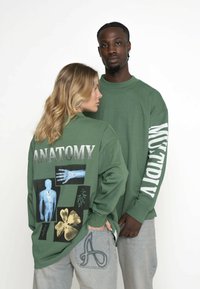 Oliven grøn langærmet skjorte med "ANATOMY" tryk og grafik af en mannequin, plante og blomst. Løs pasform med ribbede manchetter.