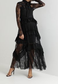 Abito in pizzo nero con maniche lunghe, strati arricciati e dettaglio a fiocco al collo. Tessuto trasparente con lunghezze variabili e una silhouette fluente.