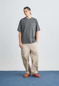 Szürke pamutpóló mellzsebbel, bézs cargo nadrággal, oldalzsebekkel, narancssárga cipőkkel, kék szőnyegen.