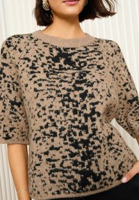 Jersey de punto en color beige con un patrón abstracto en negro, mangas cortas, cuello redondo y textura suave. Los detalles incluyen un cuello acanalado.