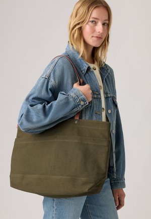 Vrouw in een lichte spijkerjack en spijkerbroek draagt een grote olijfgroene totebag met bruine handvatten over haar schouder.