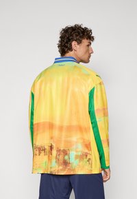 Giacca a maniche lunghe di un brillante giallo con accenti verdi. Presenta una texture liscia e design astratti, rifinita con un colletto blu.