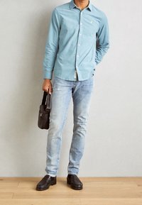 Camicia azzurra chiara con colletto, abbinata a jeans blu sbiaditi. Borsa a spalla nera e mocassini scuri. Design minimalista, look casual.