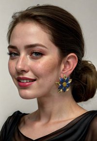 Gold flower-shaped earrings with blue accent petals and a yellow center. Textured design, featuring pointed golden accents, worn by a model.