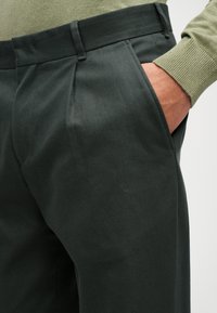 Tamnozeleni hlače izrađene od teksturiranog materijala s suptilnim dijagonalnim linijama. Imaju bočne džepove i kroj koji se prilagođava tijelu s nabranim detaljima.