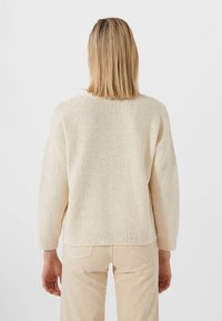 Kobieta z włosami do ramion w kolorze blond, ubrana w kremowy sweter o fakturze dzianiny i beżowe spodnie, stojąca tyłem na tle jednolitym.