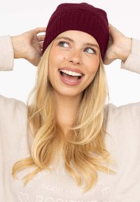 Knit burgundy beanie with textured ribbed pattern. Model has long blonde hair and wears a light sweater with embossed text.