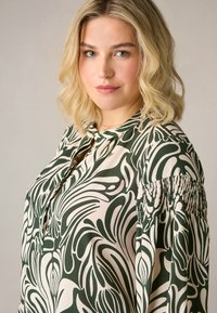 Blusa verde e crema con un motivo a foglie, caratterizzata da un colletto con apertura a chiave e dettagli di maniche arricciate, realizzata in un tessuto morbido.