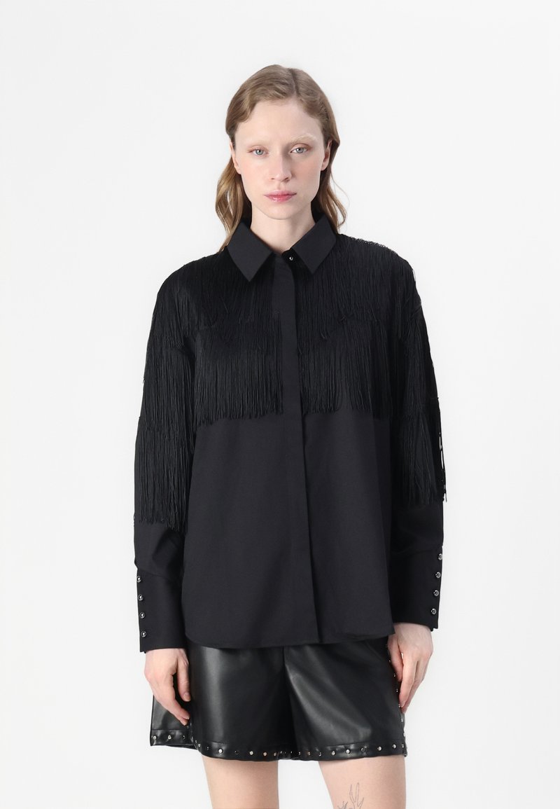 Camisa preta com textura franjada nos ombros, punhos abotoados com tachas, combinada com calções de couro pretos.