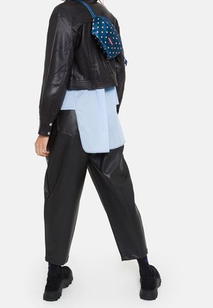 Giacca corta in pelle nera sopra una camicia azzurro chiaro con retro prolungato, abbinata a pantaloni larghi in pelle nera e sneakers nere. Piccola borsa a pois.