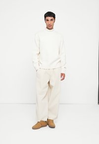 Suéter blanco de punto con cuello alto, pantalones beige de pierna ancha y zapatillas marrones. Diseño minimalista con puños y cinturilla acanalados.